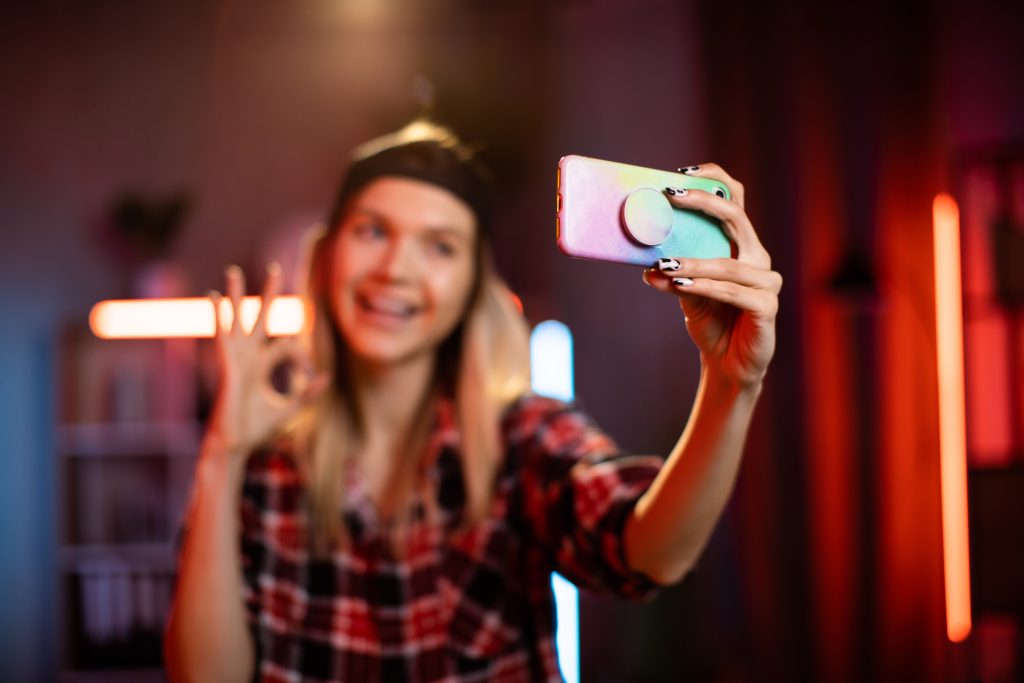 Adorable young woman wearing cap using modern smartphone for taking selfie at home. Focus on modern smartphone in female hand. Pretty female sitting at living room with evening lights with sign ok