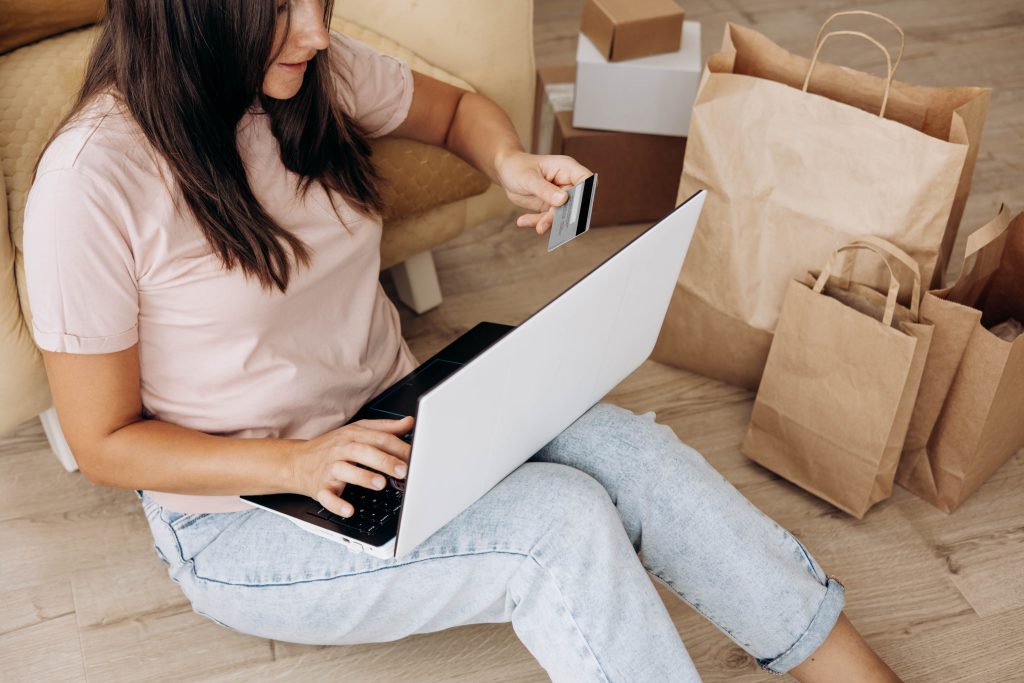 An adult woman with long brown hair sits cross-legged on a wood floor using a laptop and holding a credit card. She wears a pink t-shirt and light blue jeans. Several tan paper shopping bags and cardboard boxes sit behind her. The overall mood is light-hearted. This photo could be used for projects needing a realistic and bright shot of online shopping, financial planning, or working from home.